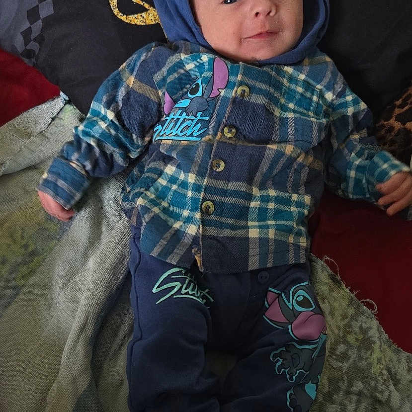 Ezio participe au concours pour gagner de l'argent avec cette photo : baby, bed, blanket, blue, child, clothing, cozy, cute, face, hood, indoor, infant, lying_down, pants, person, pillow, plaid_shirt, smile, socks, stitch_character