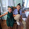 children, girls, smiling, portrait, indoor, living_room, chair, sequin_dress, green_dress, hair_bows, braids, ponytail, casual_clothing, socks, wooden_floor, windows, curtains, natural_light, family, home