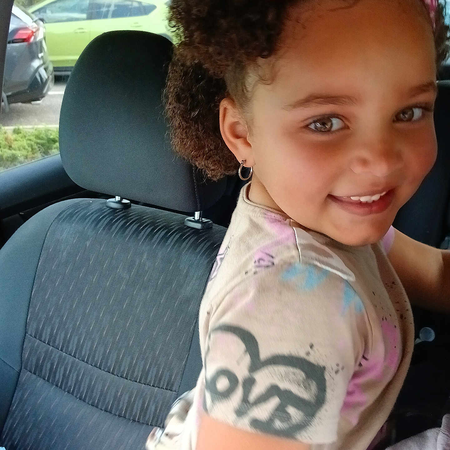 Delilah is registered to the contest to win money with this photo: child, girl, smiling, happy, portrait, bow, hair_accessory, curly_hair, earring, raccoon_shirt, car, back_seat, vehicle, car_interior, window, parked_cars, outdoor, daylight, close_up, road_trip