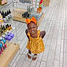 child, toddler, girl, smiling, portrait, orange_bow, floral_dress, yellow_dress, sandals, frilly_socks, earrings, standing, store, shop, retail, shelves, products, tiled_floor, indoor, fashion
