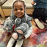 child, toddler, girl, smiling, face, braids, stuffed_animal, teddy_bear, toy, sitting, indoor, playroom, rug, colorful, chair, furniture, another_child, shoes, portrait, happy