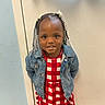 child, toddler, portrait, smiling, standing, indoor, door, hallway, denim_jacket, checkered_dress, red_clothing, braided_hair, beads, hair_accessories, fashion, looking_at_camera, dark_skin, woodwork, wall, full_body