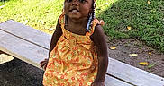 Cherish is registered to the contest to win money with this photo: beads, bench, braids, child, dress, fence, girl, grass, happy, leaves, orange_bow, outdoor, playground, playground_equipment, portrait, shoes, sitting, smiling, sunlight, toddler