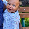baby, child, smiling, portrait, outdoor, sports_field, baseball, baseball_diamond, held, infant, face, eyes, curly_hair, earring, dress, patterned_clothing, adult, arm, wooden_bench, happy