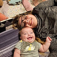 Elizabeth is registered to the contest to win money with this photo: baby, child, smiling, glasses, striped_clothing, bed, blanket, plush_toy, happy, face, person, indoors, sleeping_area, cuddly, cozy, sibling, family, cute, joy, toddler