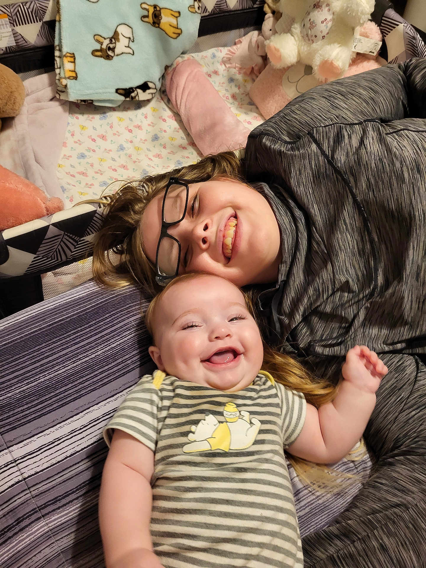 Elizabeth is registered to the contest to win money with this photo: baby, child, smiling, glasses, striped_clothing, bed, blanket, plush_toy, happy, face, person, indoors, sleeping_area, cuddly, cozy, sibling, family, cute, joy, toddler