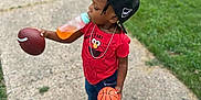 Jase is registered to the contest to win money with this photo: toddler, child, cap, bottle, football, basketball, red_shirt, elmo, chain_necklace, sidewalk, grass, car, white_shoes, jeans, outdoor, person, playing, multitasking, toy_balls, casual_clothing