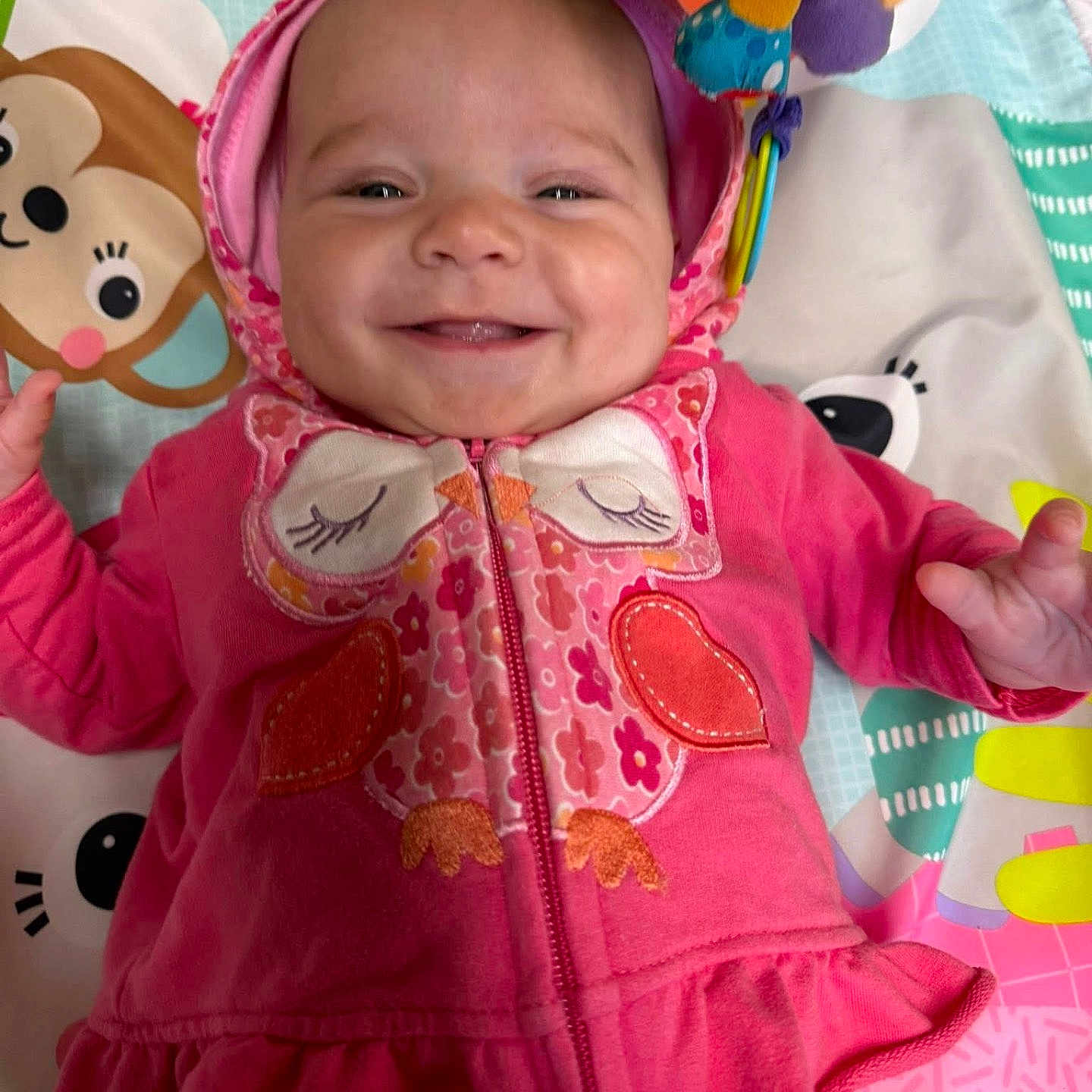 Marti joined the competition — help win amazing prizes! apparel, baby, cheerful, child, closeup, colorful, cute, face, happy, hoodie, indoors, infant, lying_down, owl_design, patterned_clothing, pink_clothing, play_mat, smiling, toy, young_child