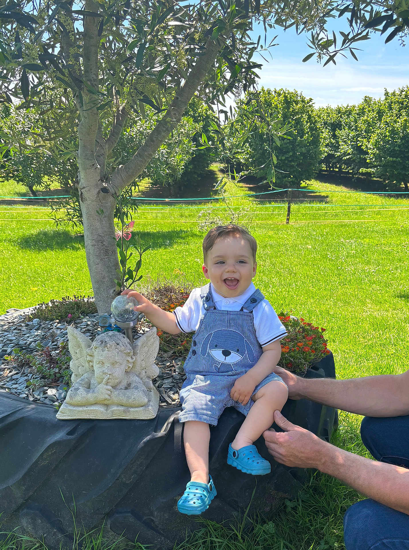 Loeven participe au concours pour gagner de l'argent avec cette photo : angel_statue, blue_shoes, child, daylight, flowerbed, garden, grass, hands, happy, nature, outdoor, overalls, plant, playful, smiling, stone_statue, sunny, support, toddler, tree