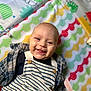 baby, smiling, child, striped_overalls, plaid_shirt, colorful_mat, patterned_mat, happy, tongue_out, cute, infant, clothing, portrait, lying_down, indoors, cheerful, face, person, playmat, closeup