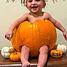 autumn, baby, bare_arms, bare_feet, child, decorations, family_photo, festive, giant_pumpkin, halloween, happy, indoor, orange_hat, portrait, pumpkin, seasonal, smiling, still_life, wall, wooden_surface