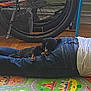 dog, person, jeans, white_shirt, bicycle_wheel, floor_mat, wooden_floor, indoor, resting, sleeping, black_dog, paw, chair, colorful_mat, toy_mat, relaxation, casual, companion, napping, animal