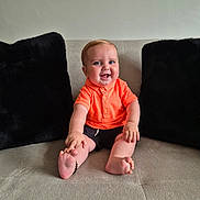 Cass is registered to the contest to win money with this photo: baby, child, smiling, orange_shirt, shorts, sofa, cushions, indoor, happy, sitting, barefoot, cute, portrait, infant, person, furniture, home, comfort, playful, young