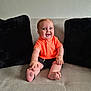 baby, child, smiling, orange_shirt, shorts, sofa, cushions, indoor, happy, sitting, barefoot, cute, portrait, infant, person, furniture, home, comfort, playful, young