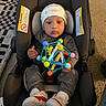 baby, car_seat, toy, hat, shoes, clothing, floor, carpet, indoor, child, infant, person, footwear, geometric_toy, pattern, headwear, cute, seated, looking_at_camera, home