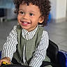 child, toddler, smiling, curly_hair, toy_car, steering_wheel, indoor, happy, clothing, striped_shirt, vest, person, play, cute, seatbelt, floor_tiles, portrait, young, joyful, kid