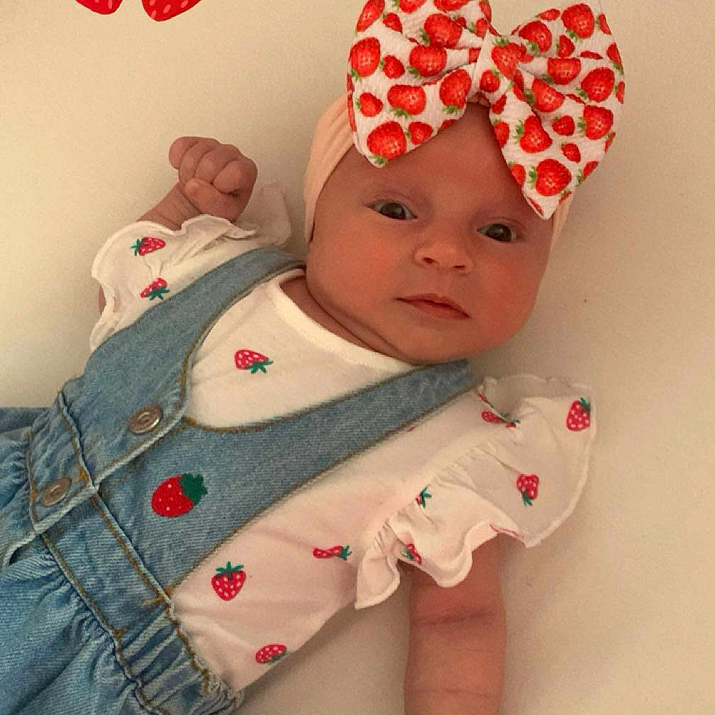 Kalyssa participe au concours pour gagner de l'argent avec cette photo : adorable, baby, bed, bow, child, clothing, cute, denim, face, fist, hand, headband, infant, lying_down, onesie, playful, portrait, soft_surface, sticker, strawberry