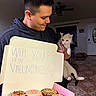man, cat, donuts, box, handwritten_sign, indoor, floor_tiles, ceiling_fan, smiling, black_hoodie, white_cat, pink_donut, brown_donut, cookie, love_message, valentines, holding, portrait, home, affection
