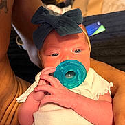 Ahzara Ourlanii is registered to the contest to win money with this photo: baby, infant, pacifier, blue_bow, white_onesie, hand, tattoo, adult_arm, closeup, indoor, person, newborn, cute, holding, skin, face, eyes, fingers, clothing, portrait