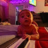baby, bed, blanket, child, clothing, curious, face, furniture, indoor, lighting, person, portrait, reaching, room, striped, television, toddler, toy, wall, young