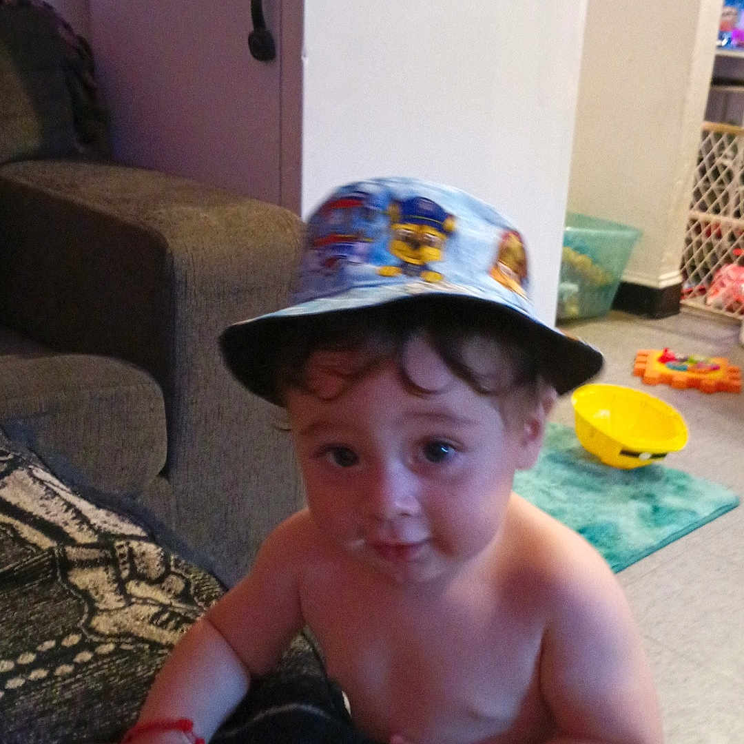 Raiden is registered to the contest to win money with this photo: baby_gate, blanket, bucket_hat, child, couch, curious, diaper, expression, face, floor, furniture, hand, hat, home, indoor, living_room, person, refrigerator, toddler, toy