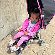 Harmony is registered to the contest to win money with this photo: baby, casual_wear, child, concrete_wall, curly_hair, cute, daylight, hoodie, outdoor, pacifier, pants, person, pink_clothing, seated, sidewalk, sneakers, stroller, sunlight, toddler, urban