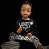 Harmony joined the competition — help win amazing prizes! black_background, casual_clothing, child, curious_expression, face, floral_pattern, hair_buns, indoor, isolated, leggings, person, portrait, shoe, sitting, snack, studio_style, sweatshirt, text_on_clothing, toddler, young_child