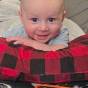 Bruce is registered to the contest to win money with this photo: baby, child, smile, blue_eyes, red_and_black_checkered, pillow, blanket, indoor, person, cute, happy, face, head, hand, toy, adult, background, relaxed, cozy, playful