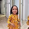 child, toddler, girl, smiling, happy, portrait, standing, floral_dress, yellow_clothing, curly_hair, indoors, hallway, mirror, reflection, closet, door, tile_floor, home, fashion, photography