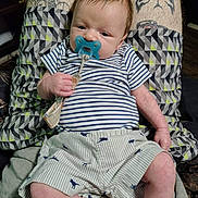 Jameson joined the competition — help win amazing prizes! baby, pacifier, person, tattoos, shorts, striped_shirt, dinosaur_print, legs, hand, face, indoor, clothing, pattern, holding, infant, cute, relaxed, skin, seated, comfort