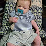 baby, pacifier, person, tattoos, shorts, striped_shirt, dinosaur_print, legs, hand, face, indoor, clothing, pattern, holding, infant, cute, relaxed, skin, seated, comfort