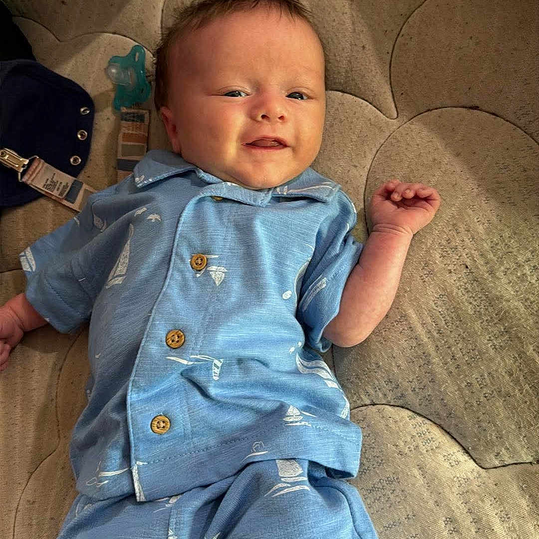 Jameson is registered to the contest to win money with this photo: baby, infant, blue_clothing, smiling, lying_down, hand, indoors, fabric, patterned_clothing, sailboat_pattern, cute, child, person, short_sleeves, shorts, soft_surface, comfort, skin, head, face