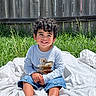 Santiago joined the competition — help win amazing prizes! child, toddler, smiling, curly_hair, blue_shirt, denim_shorts, blanket, grass, outdoor, sunlight, fence, happy, sitting, portrait, nature, daylight, casual_clothing, cute, playful, young