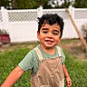 child, toddler, smiling, curly_hair, overalls, green_grass, backyard, white_fence, outdoor, happy, cute, young_child, portrait, casual_clothing, daylight, person, nature, playful, standing, boy