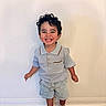 Santiago is registered to the contest to win money with this photo: child, toddler, smiling, curly_hair, standing, light_blue_shirt, denim_shorts, brown_shoes, indoor, wooden_floor, plain_wall, happy, cute, young_child, casual_clothing, portrait, small_person, feet, hands, bright