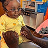 Brooklyn is registered to the contest to win money with this photo: toddler, child, glasses, yellow_pajamas, holding_hands, adult, playroom, toy_kitchen, calendar, indoor, smiling, braided_hair, black_skin, person, furniture, colorful, curtains, floor, seat, home