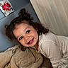 Aniyah joined the competition — help win amazing prizes! child, toddler, smiling, curly_hair, blanket, bedroom, white_clothing, indoor, happy, person, face, blue_eyes, hair_buns, soft_texture, cushion, wall, pillow, cozy, young, playful