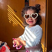 Aniyah is registered to the contest to win money with this photo: toddler, child, sunglasses, heart_shaped, pink, white_shirt, purple_skirt, indoor, sunlight, shadow, wooden_door, clothing_pile, carpet, smiling, curly_hair, happy, playful, person, portrait, cute