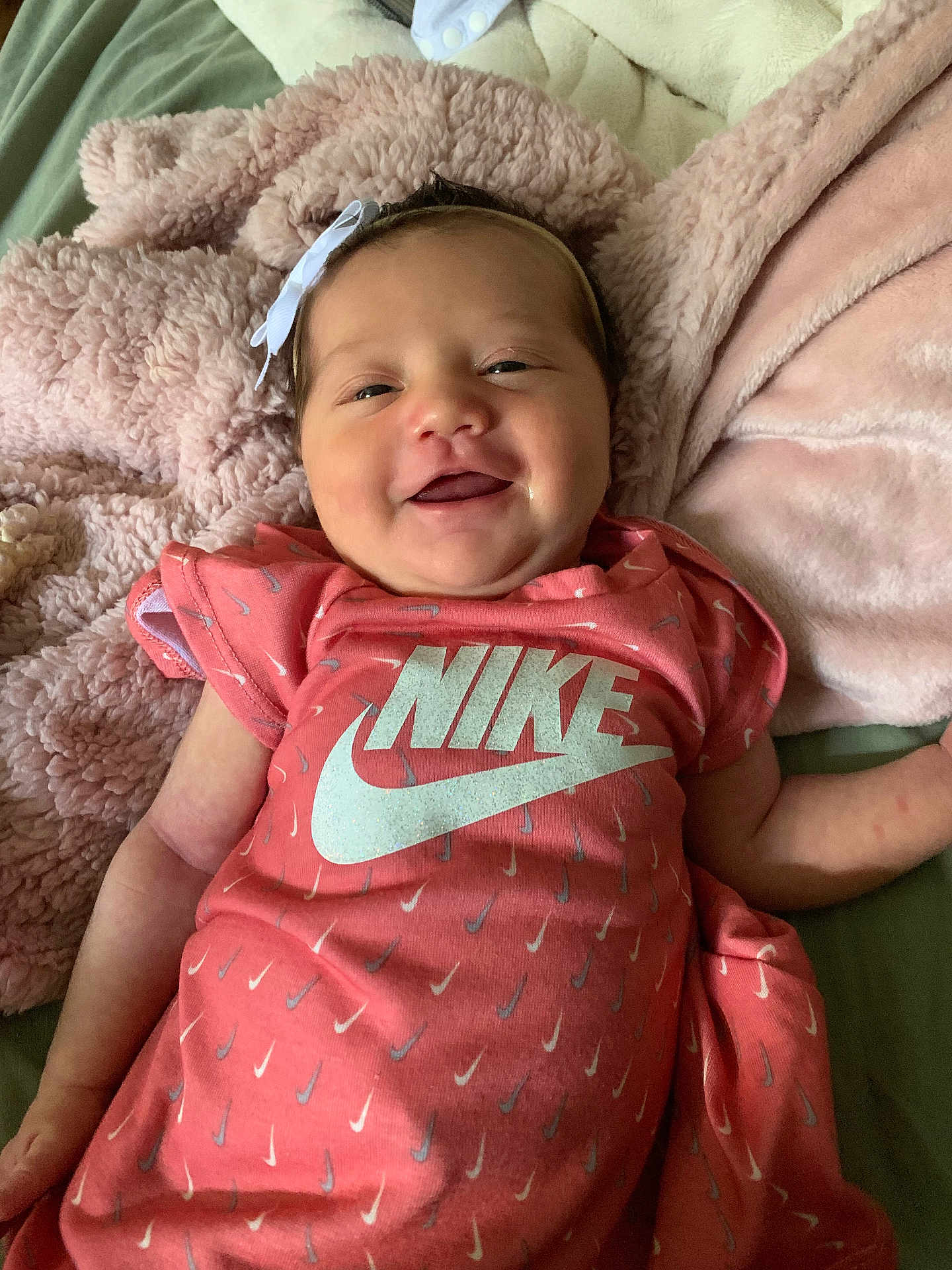 Mariana is registered to the contest to win money with this photo: baby, infant, smile, pink, nike, onesie, bow, blanket, soft, happy, cute, child, face, clothing, apparel, person, portrait, lying_down, indoors, cozy