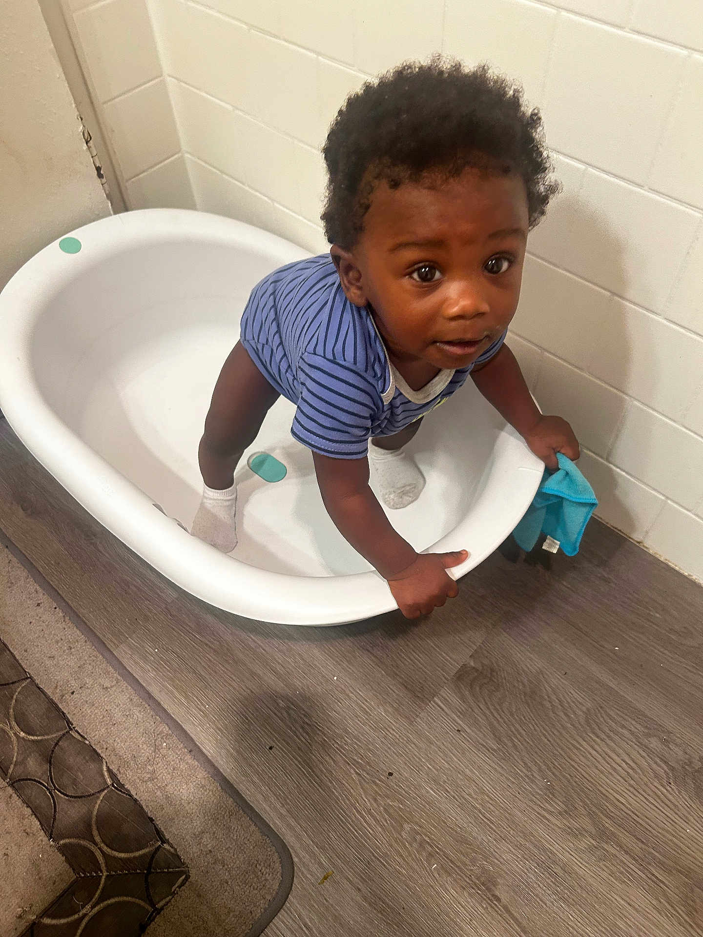 James is registered to the contest to win money with this photo: toddler, child, baby, onesie, striped_clothing, plastic_tub, floor, wood_floor, tiled_wall, socks, blue_cloth, indoor, standing, holding, curious, person, young_child, baby_clothing, grip, looking_up