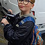 child, glasses, scooter, backpack, black_jacket, sidewalk, urban, van, outdoor, short_hair, person, smirk, street, vehicle, hoodie, hand, casual_clothing, daylight, curb, young
