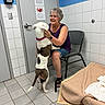 affection, animal_shelter, blanket, chair, collar, dog, door, glasses, hand, happy, indoor, pet, pet_bed, shorts, smile, tile_floor, tile_wall, veterinary_clinic, watch, woman