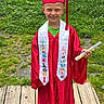 child, young_graduate, graduation, cap, gown, diploma, smiling, portrait, outdoor, grass, flowers, wooden_bridge, school, ceremony, red_clothing, white_stole, tassel, standing, happy, academic_achievement