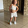 bottle, carpet, casual, child, door, expression, girl, hands_on_hips, indoor, knitted_clothing, orange_liquid, pigtails, pose, reflection, stone_wall, tiled_floor, white_shoes, white_shorts, white_top, young_child