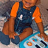 Makhii is registered to the contest to win money with this photo: toddler, child, smiling, toy, piano, blue, orange_clothing, denim_overalls, indoor, bed, pillow, blanket, happy, person, cute, playing, baby, sitting, face, cozy