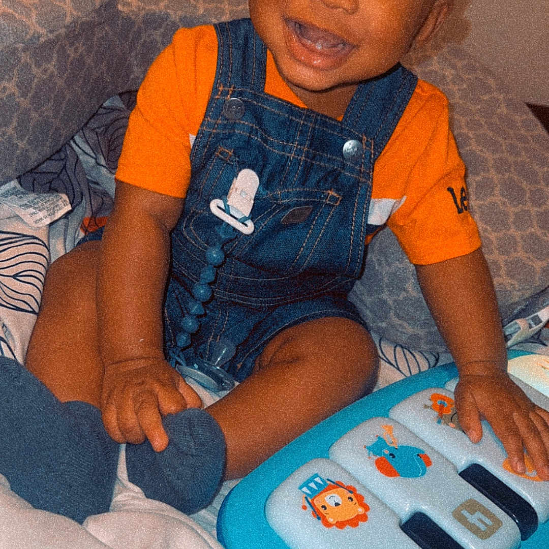 Makhii is registered to the contest to win money with this photo: toddler, child, smiling, toy, piano, blue, orange_clothing, denim_overalls, indoor, bed, pillow, blanket, happy, person, cute, playing, baby, sitting, face, cozy