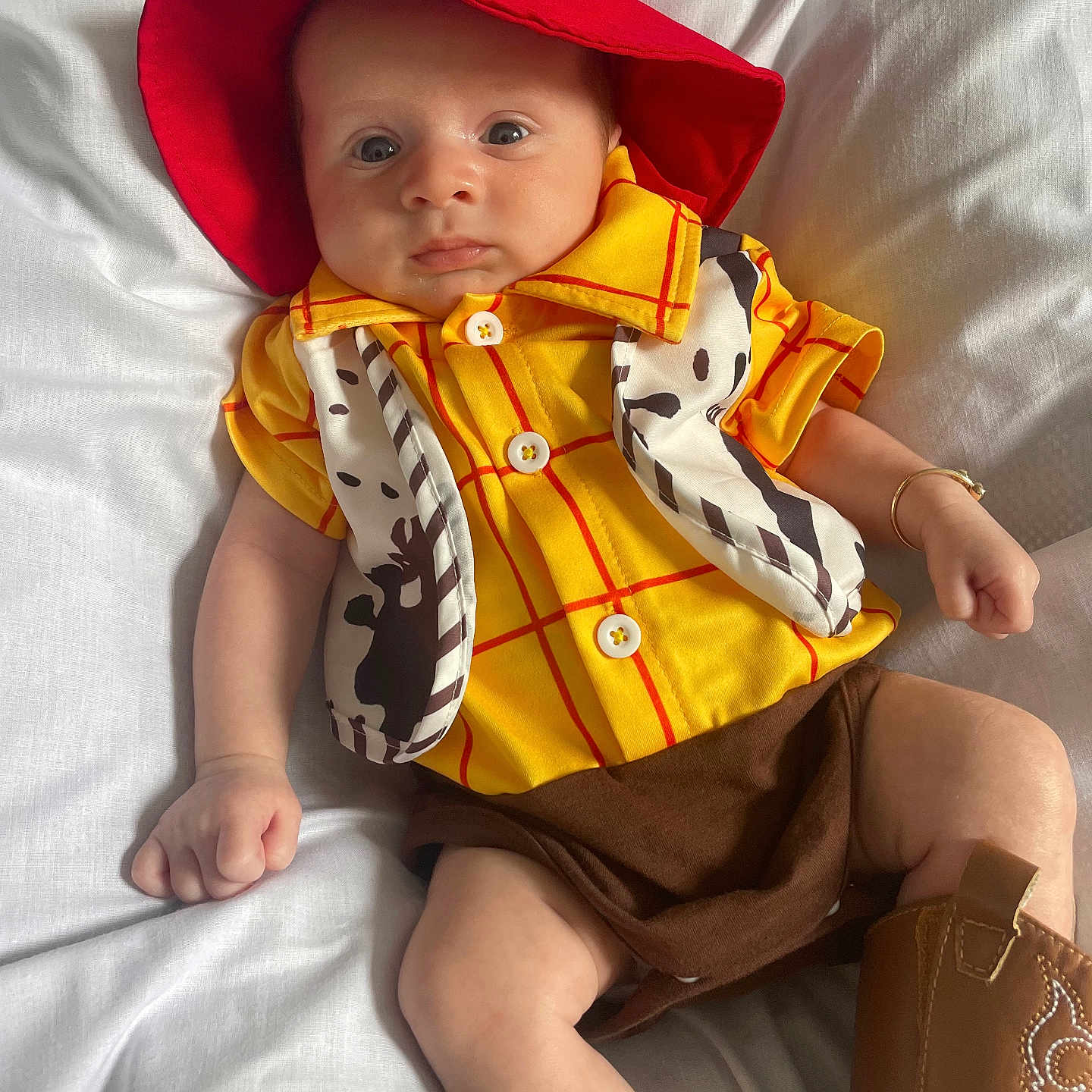 Bobby Lee is registered to the contest to win money with this photo: accessory, baby, brown_shorts, child, costume, cow_print_vest, cowboy_boots, cowboy_costume, cute, fashion, footwear, hat, indoors, infant, lying_down, person, portrait, red_hat, white_blanket, yellow_shirt