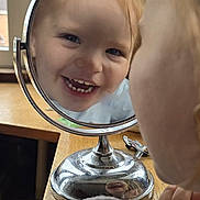 Lottie-Jae is registered to the contest to win money with this photo: toddler, child, mirror, reflection, smile, happy, face, teeth, wooden_table, window, indoor, portrait, close_up, person, furniture, light, hair, hand, cozy, clothing