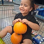 Anthony is registered to the contest to win money with this photo: child, toddler, smiling, grocery_store, shopping_cart, pumpkin, autumn, harvest, black_clothing, curly_hair, portrait, sitting, indoor, supermarket, food, snacks, cart, happy, family_shopping, seasonal