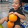 child, toddler, smiling, grocery_store, shopping_cart, pumpkin, autumn, harvest, black_clothing, curly_hair, portrait, sitting, indoor, supermarket, food, snacks, cart, happy, family_shopping, seasonal